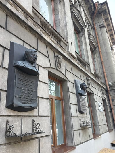 This media file is a photograph taken from a low angle looking up at the facade of a building. The building appears to be made of stone tiles and has multiple windows. There are two prominent memorial plaques on the facade.

The lower plaque features a bronze bust of a man, identified by the inscription as "АНАТОЛИЙ ИВАНОВИЧ ПОТАПОВ". The inscription also states he was an "АКАДЕМИК РАМН" (Academician of RAMS), "ОСНОВАТЕЛЬ И ПЕРВЫЙ ДИРЕКТОР" (Founder and First Director) of the "НИИ ОНКОЛОГТА" (Research Institute of Oncology), and provides the years of his life as "1935-2013". The text on the plaque is in Cyrillic script.

Above this plaque, there is another smaller plaque with a bust of a person. There are also decorative architectural elements on the building's facade, including ornate metal brackets and carved stone faces. A copper-colored downspout runs along the upper portion of the building. The overall scene suggests a historical or significant building, likely in an urban setting given the architecture. The lighting suggests it is daytime. The location context provided is Tomsk, Russia.