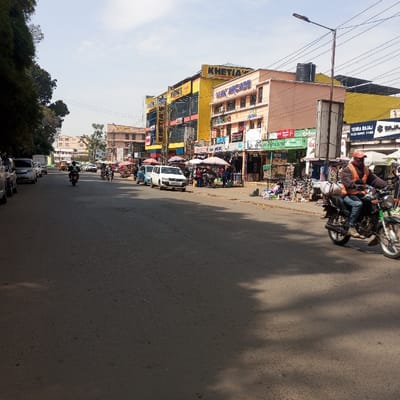 The image displays an urban street scene in Sheywe ward, Kenya, under clear daylight conditions, indicated by strong shadows. The paved asphalt road, showing minor surface cracks, occupies the foreground and central mid-ground.
On the right side of the road, a male individual is operating a dark-colored motorcycle, moving towards the right frame. He is wearing an orange safety vest, blue jeans, and a red cap. A white cylindrical object is affixed to the rear of the motorcycle. Along the right curb, several commercial buildings are visible. A prominent yellow, multi-story building features "KHETIA'S" and "LINK ARCADE" signage. Adjacent to it is a lighter-colored, multi-story building. The ground level of these buildings contains various storefronts. A series of street vendors operate along the sidewalk, utilizing multiple white canopy tents to display merchandise, including clothing and other goods, on the ground and on tables. Several pedestrians are present in this area, some appearing to interact with the vendor stalls. A white van is parked parallel to the curb on the right. Overhead utility lines and a street lamp pole are also visible on the right.
On the left side of the road, a line of parked vehicles, including white vans and sedans, is visible alongside green foliage. Further down the street in the background, additional vehicles, including motorcycles and cars, are present, indicating ongoing traffic. A few other motorcyclists are visible further down the road, some with passengers.