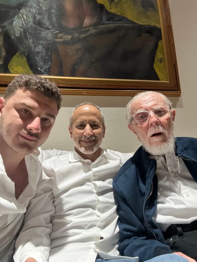 Three men pose in front of a large framed painting. The oldest man, with white hair, beard, and glasses, is at the top, wearing a blue jacket over a white shirt. A man with a shaved head and a white shirt is in the middle. The youngest man with short brown hair and a white shirt is in the foreground. They are all smiling, and the image has a slightly tilted perspective. The painting is mostly dark, but parts appear yellowish.