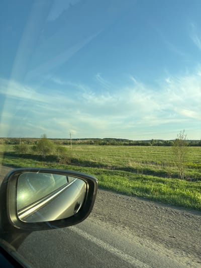 The image depicts a view from inside a vehicle, likely on a road trip. The scene outside the car features a clear blue sky with light clouds. A grassy field extends along the roadside, with trees visible in the distance. A gray side-view mirror is visible, reflecting light and part of the car's interior. The road is asphalt and appears to be in decent condition. The location is in or near the city of Shavkunovo, Russia. There are no people visible.