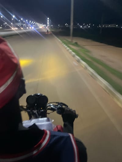This media file depicts a first-person perspective of someone riding a motorcycle at night in Gbessa, Nigeria. The rider is wearing gloves and a dark jacket with red and white stripes. The motorcycle's headlights illuminate a road with a light-colored surface and a green striped border on the left side. In the distance, a line of streetlights stretches along the road, indicating an urban or semi-urban setting. The sky is dark, suggesting it is nighttime. The overall impression is one of movement and travel through a dimly lit environment. There is no visible text in the image.