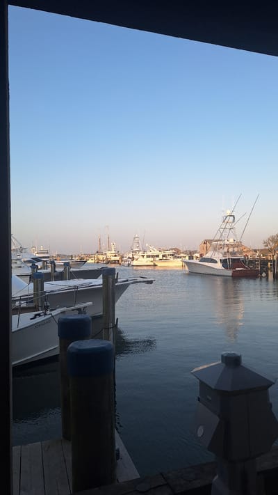 This image shows a view of a harbor in Nantucket, Massachusetts, United States of America. The scene is framed by a dark border, suggesting it was taken from inside a building or under an overhang. The water is calm, reflecting the clear blue sky and the boats docked in the harbor. Several large yachts and fishing boats are visible, with masts and rigging extending upwards. In the foreground, wooden pilings with blue caps are present on a dock. The lighting suggests it is either early morning or late afternoon, with the sun casting a warm glow on the scene. The overall atmosphere is tranquil and serene, characteristic of a harbor setting.