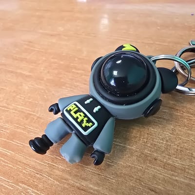 This is a close-up, eye-level shot of a keychain featuring a small, stylized astronaut figurine. The figurine is predominantly gray with black accents, including a large, reflective black visor and two small black ear-like protrusions on its head. The astronaut is wearing a black and gray suit with a bright yellow rectangle on its chest that has the word "PLAY" written in black, pixelated letters. The figurine is attached to a metal keychain with several interlocking silver rings. The background is a textured, light brown wooden surface, possibly a table or desk, with visible grain and vertical lines. The lighting suggests an indoor setting with ambient light, as there are reflections on the astronaut's visor and a slight sheen on the wooden surface. There are no discernible indicators of the time of day, weather, or specific emotions. The location context provided is Bryansk, Russia, but this is not visually evident from the image itself.