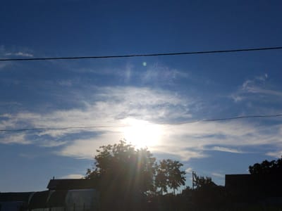This is a full shot of a bright, sunlit sky above a rural landscape in Cîrpești, Moldova. The sun is prominently visible, positioned in the upper-middle section of the frame, creating a lens flare effect with visible light rays. Scattered clouds, some appearing wispy and others more dense, streak across the deep blue sky. Two horizontal power lines traverse the sky, one in the upper third and another closer to the middle. In the foreground, silhouetted against the bright sky, are the dark shapes of trees and structures, including what appear to be greenhouses or sheds with arched tops, and a dark-roofed building. The overall impression is of an outdoor scene, likely taken during daylight hours in a rural setting.