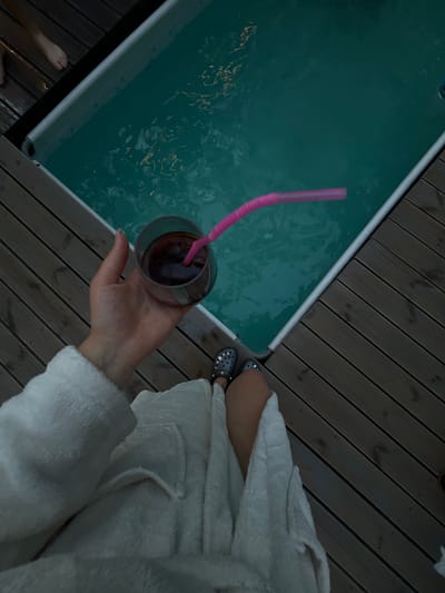 The image depicts a personal perspective of a person wearing a white bathrobe, holding a glass of dark liquid with a pink straw. The person is sitting on a wooden deck next to a small pool. The pool water is a turquoise color, and ripples are visible on its surface, reflecting light. The deck is constructed from wooden planks arranged diagonally. In the upper left corner of the image, a person's bare feet are visible on the deck, suggesting another individual is present. The scene appears to be outdoors, possibly in a backyard or relaxation area. The image does not contain any information that places the scene specifically in Ivachiv Horishnii, Ukraine.