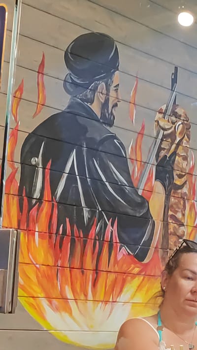 The image depicts a mural painted on a wooden wall, likely at a food establishment. The mural features a man in a dark turban and coat, seen from the back, tending to a vertical spit of meat with flames licking around him. To the right of the mural, the shoulder and head of a woman wearing a turquoise top and sunglasses are visible. The woman appears to be looking down or to the side. The overall setting suggests an outdoor or semi-outdoor location, possibly a restaurant or cafe. The style of the mural, with its depiction of a food preparation scene and vibrant flames, suggests it's advertising a type of cuisine. There is no specific indication of the city of Dagomys, Russia within the image itself.