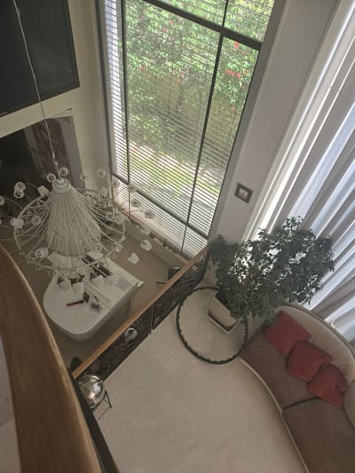 This is a high-angle, wide shot of a spacious, well-lit living room in Kfar Saba, Israel. The room features a double-height ceiling with large windows that overlook lush greenery outside. A large, intricate white chandelier hangs prominently in the center of the space. Below it, a white grand piano sits with a red pen and a small framed picture on its surface. A dark metal railing with ornate scrollwork lines a staircase. To the right, a large potted plant with dense green foliage stands next to a curved sofa adorned with three red throw pillows. Light-colored tiled flooring covers the ground. The overall impression is one of modern elegance and comfort, with a strong connection to the natural surroundings.