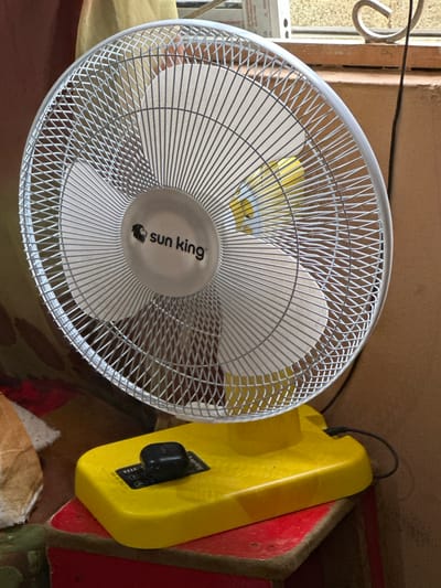An up-close, medium shot shows a white wire grill surrounding the blades of a fan. The fan has a circular, silver center with the "sun king" logo visible. The fan's base is bright yellow plastic, with a black button on top, and it rests on a red surface. The background is a blur of warm tones, suggesting an indoor setting. The image is likely a product photograph or a personal snapshot of the fan. The city of Gidan Waziri, Nigeria is not visible in this image.