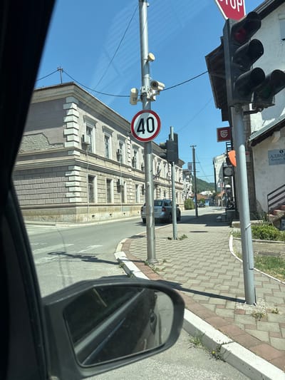 The image depicts a street scene in the city of Zvornik, Bosnia and Herzegovina, viewed from inside a vehicle. The perspective is primarily from the driver's seat, as indicated by the presence of a side mirror in the lower left of the frame.

In the foreground, the road surface is visible, with a pedestrian crossing marked by white lines. A gray sedan is parked on the right side of the road, facing away from the viewer. Adjacent to the road is a paved sidewalk made of interlocking bricks.

Dominating the center of the image is a utility pole with various fixtures attached. These include a traffic light with red and green signals illuminated, a speed limit sign indicating "40" within a red circle, and what appear to be security cameras. Further down the street, another utility pole is visible.

On the left side of the street, a large, ornate building with classical architectural elements stands prominently. It has multiple windows and air conditioning units installed. The building's facade is light-colored with decorative stone detailing.

The environment is bathed in bright sunlight, suggesting a clear day. The sky is a vibrant blue. In the background, the street continues, lined with more buildings and vegetation, with hills visible in the distance. The overall impression is that of a typical urban street in a European city.