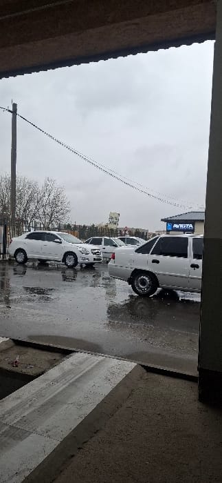 The image, when oriented correctly, displays an outdoor street or parking lot scene on an overcast day. The ground is dark, wet, and reflective, suggesting recent rain or damp conditions, with visible puddles.
The primary subjects are several white cars, mostly sedans or small utility vehicles, parked in a line facing to the right of the frame. Their uniform white color stands out against the muted tones of the environment. In the immediate foreground on the lower right, concrete barriers or structures are visible.
To the left, a building with a textured brown wall is partially visible, while further back, bare trees line the street, indicating a colder season. Utility wires crisscross the upper part of the sky, and a utility pole is present on the left side. In the background, above the parked cars, a building features a prominent blue sign. The text on this sign is clearly visible and reads "AVISTA" in white letters. The sky is a uniform, light grey, completely overcast, providing a diffused and somber light to the scene.
There are no people or active events taking place; the scene depicts static parked vehicles. The overall atmosphere is neutral, characterized by the grey weather and damp conditions. The location is Khalkabad, Uzbekistan, as per context.
