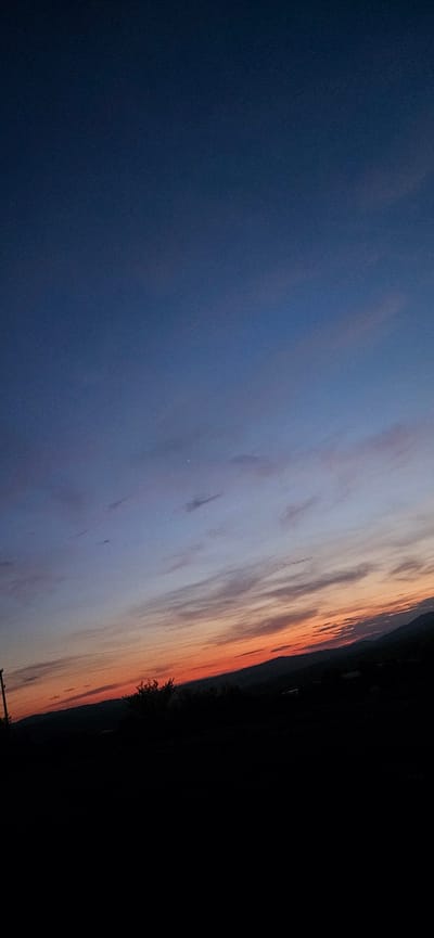 The image depicts a sunset over a rural landscape, likely Momin Prohod, Bulgaria. The sky transitions from a deep navy blue at the top to lighter blues and then streaks of orange and pink near the horizon. Thin, wispy clouds are scattered across the sky. Silhouetted against the sunset is a dark, undulating treeline and hills. In the foreground, a dark, silhouetted object, possibly a utility pole, is visible on the left side. The bottom portion of the image is entirely black, obscuring any ground-level details. There are no people or visible actions taking place.