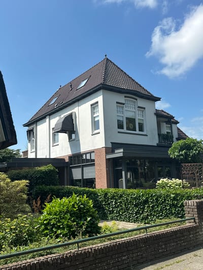 A two-story white stucco house with a dark tiled roof and dormer windows is situated in a suburban environment under a clear blue sky with scattered white clouds. The house features multiple windows, including a large bay window on the second story with a decorative transom. A dark retractable awning is extended over a ground-floor window. The ground level of the house incorporates a section with red brickwork and large glass windows, suggesting a conservatory or extension. Lush green vegetation, including hedges and shrubs, surrounds the property, with a brick wall and a green metal railing in the foreground. The scene is likely captured on a sunny day.