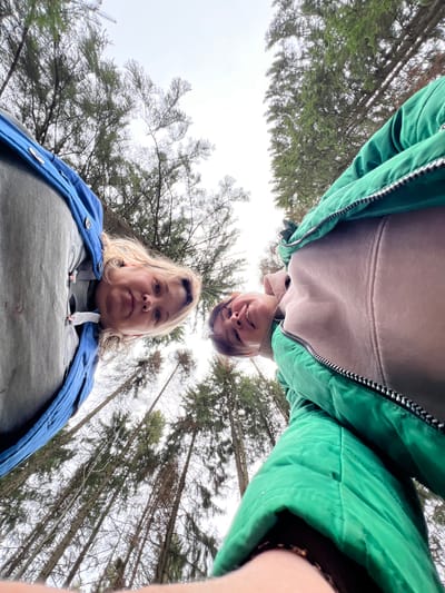 The image depicts two individuals standing outdoors in a wooded area. The perspective is from below, looking upwards. The individual in the foreground is a woman with blonde hair, wearing a gray shirt and a blue jacket. The second individual, above and slightly behind her, appears to be wearing a light pink hoodie and a green jacket. Trees surround the subjects, reaching towards the sky. The weather appears overcast. The exact location is Bol'shakino, Russia. There are no visible buildings or other man-made structures. The interactions between the people are indistinguishable.