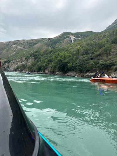 The image depicts a scene on a turquoise river, likely the Chirkey Reservoir in Chirkey, Russia, with a steep, vegetated mountainside forming the backdrop. In the foreground, the dark hull of a watercraft occupies the left side of the frame, angled towards the water. Further into the river, an orange speed boat with a black canopy is visible, carrying at least three individuals. The boat has the text "GGG" prominently displayed on its side. Another smaller boat, possibly a sailboat, is further upstream on the left side of the river. The water exhibits ripples and is a vibrant, clear green. The sky above is overcast, casting a diffused light on the landscape.