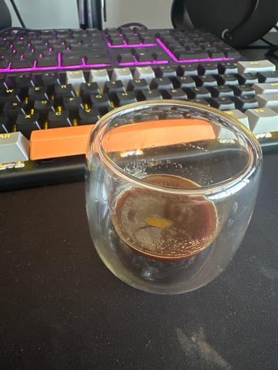 The scene depicts a mechanical keyboard with black, gray, and orange keycaps, illuminated with purple LED backlighting. A clear glass containing a murky brown liquid with bubbles sits atop the keyboard. The setting is a desk covered with a black mousepad. There are no people visible in the image. The location is not deducible from the image.