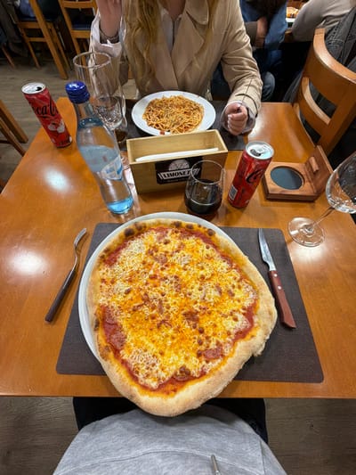 The image captures an overhead view of a dining table at Limonet Esplanada & Pizzaria, located in Peso da Régua, Portugal. In the foreground, a large, round cheese pizza, with a thick crust and uniform golden-melted cheese over a red tomato base, rests on a white plate centered on a dark placemat. A silver fork with a dark brown handle lies to the left of the pizza, and a matching knife is to its right. To the upper left of the pizza, on the bare wooden table, is a closed, light blue plastic bottle of water next to a tall, empty clear glass. Adjacent to these is a red Coca-Cola can featuring a white Santa Claus graphic. Further right, a wooden napkin holder displaying the "LIMONET Esplanada & Pizzaria" logo contains white paper napkins. A clear glass with a dark liquid (possibly red wine) is next to the napkin holder. An open, red Coca-Cola can is positioned beside the dark liquid glass, and to its right is a small, open wooden box or coaster. A clear stemmed wine glass is visible on the far right. Directly opposite the pizza, a plate of spaghetti with red sauce is positioned in front of a person with light-colored hair, wearing a light-colored coat, whose hands are resting on the table. The lower torso and lap of another person, wearing a grey hooded sweatshirt, are visible in the bottom foreground, indicating the photographer's perspective. Blurred figures and wooden chairs are discernible in the background, suggesting a bustling restaurant setting.