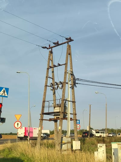 This is a medium-shot, eye-level, outdoor photograph taken in Cząstków Mazowiecki, Poland. The scene is dominated by a tall, wooden utility pole with a transformer and electrical wires. To the left of the pole, a traffic light is visible, with a pedestrian crossing sign above it. Further to the left, a street light illuminates the scene. In the background, several vehicles are on a road, including vans and cars. The area is bordered by dry grass and trees. The sky is clear blue with some wispy clouds.