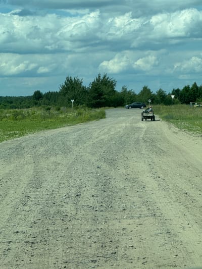 An outdoor, daytime, medium shot captures a gravel road leading into the distance, bordered by green grass and trees. A person is seated in a small, four-wheeled utility vehicle with a trailer, traveling on the road. In the background, a dark-colored car is parked on the side of the road. The sky is filled with dramatic, cloudy formations. The scene appears to be located in Orlivka, Ukraine.