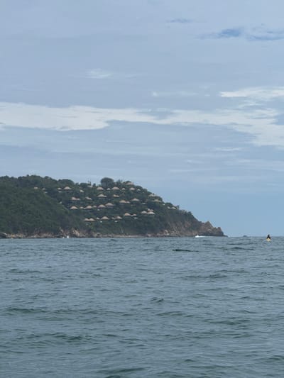 The image depicts a panoramic view of the coastline at Puerto Marqués, Mexico, under an overcast sky. The foreground is dominated by a expanse of choppy, dark blue-grey ocean water. In the midground, a verdant, densely forested hillside rises sharply from the water, covered in various shades of green foliage. Scattered across the slope of this hill are numerous structures, each featuring a distinctive reddish-brown, often domed or conical roof, indicative of a resort or residential development integrated into the natural landscape. The base of the hill along the water reveals rugged rock formations where small waves are visibly breaking.
On the far left, a yellow buoy floats near the shore. On the right side of the frame, further out in the water, a solitary individual on a bright yellow kayak or paddleboard is visible. The sky above is largely covered with an unbroken layer of light grey and white clouds, suggesting an overcast or cloudy day with soft, diffused lighting.