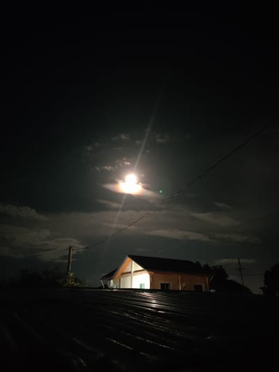 This media file is a nighttime photograph taken from a low vantage point, likely a rooftop, looking up at a house and the sky. The foreground is dominated by a dark, corrugated metal roof, its texture and ridges clearly visible due to the ambient light reflecting off it.
In the mid-ground, a two-story house is illuminated from within, casting a warm, yellowish light onto its exterior and some of its surroundings. The house has a visible roof with a pitched design, and windows are present on its facade. Trees and foliage are visible to the left of the house, silhouetted against the night sky. A wooden utility pole stands to the left of the house, with electrical wires stretching across the frame, adding to the sense of an inhabited, possibly residential, area.
The sky above is dark, indicating nighttime. A bright, luminous moon is visible, partially obscured by clouds. The moon's light creates a halo effect and a distinct lens flare, with visible light rays emanating from it. The clouds appear wispy and scattered, suggesting a partly cloudy sky. A few faint stars are also discernible in the darker areas of the sky.
The overall mood is serene and slightly mysterious, with the contrast between the dark foreground and the illuminated house and moon creating a visually striking image. The presence of utility poles and wires suggests an urban or suburban setting. The location context of San Pascual, Philippines, places this scene in a tropical or subtropical region. No text is visible in the image.