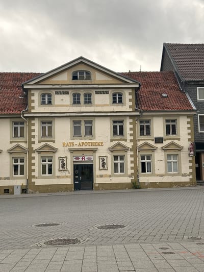The image shows the RATS-APOTHEKE building, a pharmacy, located in the city of Königslutter am Elm, Germany. The building has a light-colored facade with contrasting trim and multiple windows. Above the entrance, the name "RATS-APOTHEKE" is displayed in gold lettering. A green door leads into the pharmacy. On either side of the entrance are decorative elements that appear to be snakes coiled around poles. The building is situated on a cobblestone plaza, with several manhole covers visible. The sky is overcast. The architecture suggests a historical setting.