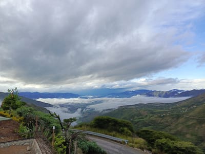 The image captures a high-altitude view in El Cisne, Ecuador. The scene is dominated by a valley filled with low-lying clouds, partially obscuring the terrain below. Mountain ranges are visible in the background, some appearing above the clouds. The foreground features a winding road with a metal guardrail. Lush green vegetation covers the slopes leading down to the valley. In the left foreground, there's construction work with reinforcing bars and newly laid ground. The sky is overcast with thick, gray clouds, though patches of blue sky are visible toward the right.