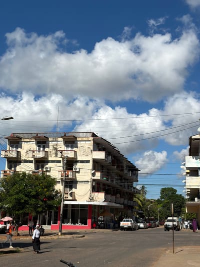 The image depicts a street scene in Pemba, Mozambique. A multi-story building occupies a significant portion of the frame, with several balconies and air conditioning units visible. Utility lines crisscross the sky in front of the building. The sky itself is filled with white cumulus clouds against a blue backdrop.

On the right side of the image, a street runs along the building. People can be seen walking on the sidewalk. Parked vehicles line the street. A leafy tree is positioned in front of the building at street level. The street surface appears paved but slightly worn. A metal pole stands at the edge of the street.