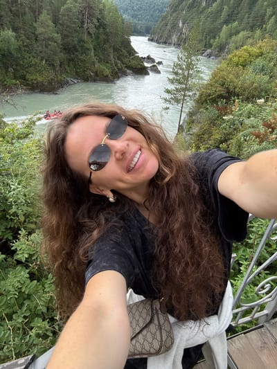In Chemal, Russia, a woman with long, curly brown hair, wearing sunglasses and a black t-shirt, is taking a selfie on a wooden platform overlooking a fast-flowing river. She has a bright smile and is wearing a pearl earring. A beige Gucci crossbody bag is slung over her shoulder, and a white hoodie is tied around her waist. In the background, a group of people in red life vests are rafting down the river, surrounded by lush green trees and steep, rocky banks. The scene captures a moment of adventure and natural beauty.