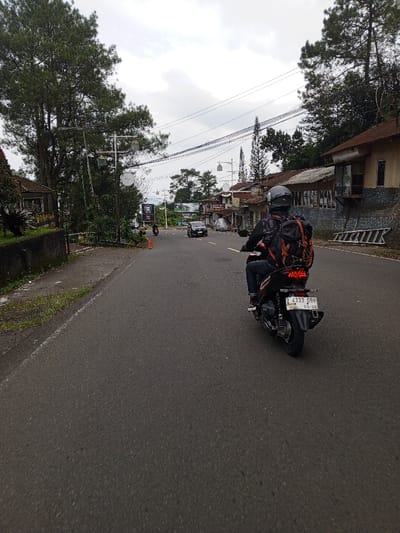 The scene depicts a rear view of an individual operating a black scooter on an asphalt road in Ciloto, Indonesia, under an overcast sky. The scooter operator wears a black full-face helmet, a dark long-sleeved garment, dark trousers, and a prominent black and orange backpack. The scooter's rear features illuminated red taillights and an Indonesian license plate, "Z 4333 DBR," with an expiration date of March 2029. The road, devoid of clear lane markings in the foreground, extends into the distance where a dark sedan and several other motorcycles are visible, some appearing to travel in the opposite direction. An orange traffic cone rests on the left shoulder of the road further ahead. To the immediate left, a concrete barrier with vegetation gives way to a tall pine tree and a multi-lamp street light. To the right of the road, a series of single to two-story buildings, consistent with commercial or residential structures, line the street, some displaying stone or brick facades. A white step ladder leans against a stone retaining wall adjacent to one of these buildings. Overhead, multiple utility poles support a network of power lines. Dense green foliage and additional pine trees are visible along both sides of the road and in the distant background.