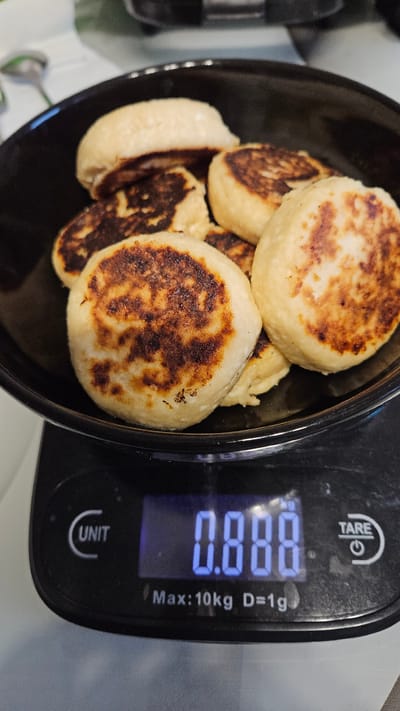The image displays a close-up, top-down view of food items being weighed on a digital kitchen scale. A dark-colored bowl filled with several round, pan-fried food items is positioned on the scale's platform. These items, resembling small pancakes or cheesecakes, have a light, doughy appearance with distinct golden-brown, crispy patches from frying.

The digital scale, black in color, shows a reading of "0.880" in blue illuminated digits. The unit "kg" is visible next to the numerical display, indicating the weight is 0.880 kilograms. Other text on the scale includes "TARE" and a power symbol, "UNIT" with a curved arrow, and technical specifications like "Max:10kg D=1g". No people are visible in the frame. The background is indistinct, suggesting an indoor setting, likely a kitchen. The time of day and specific emotions are not discernible from the image.