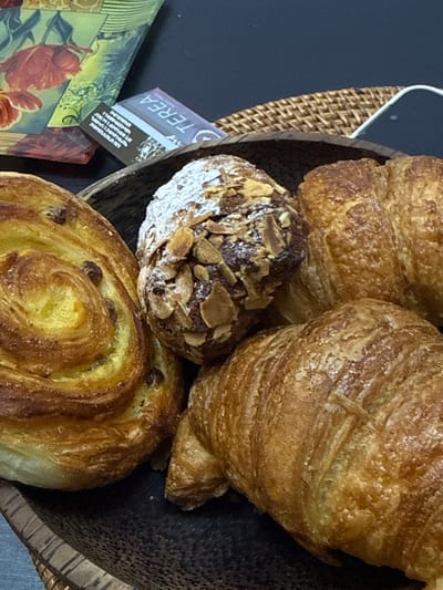 A close-up shot shows an assortment of baked goods arranged in a dark brown, woven basket, likely inside a food establishment in Zovuni, Armenia. Two croissants with flaky, golden-brown crusts occupy the upper part of the basket. Nestled among the croissants is a round pastry dusted with powdered sugar and topped with sliced almonds. In the foreground, a spiral-shaped pastry with a shiny, golden-brown surface speckled with raisins is visible. A small, rectangular object with the partially visible word "TEREA" is positioned near the edge of the basket, possibly a product packaging or a label. A small decorative item with floral elements is placed next to the basket. The background is dark and out of focus.