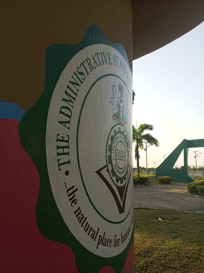 The image shows the logo of the Administrative Staff College of Nigeria (ASCON) on a pillar in the city of Tawpo, Nigeria. The logo is a circular design with the college's name surrounding a central emblem containing the letters ASCON and the words "College of Nigeria". The Nigerian coat of arms is positioned above the central emblem. The pillar is decorated with colorful paint around the logo. In the background, the college's entrance gate, palm trees, and other landscaping are visible under a bright sky.