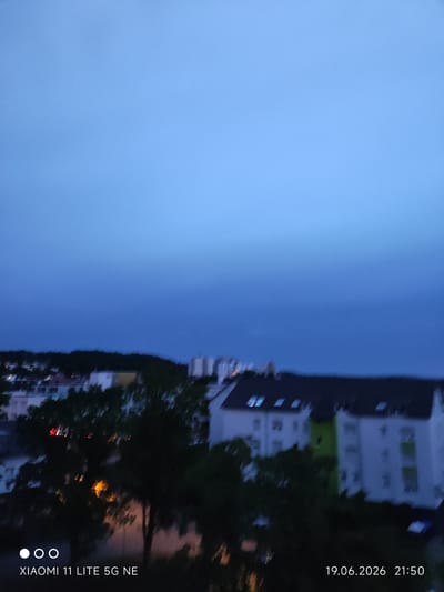 The image shows a twilight view of the city of Böblingen, Germany, from an elevated perspective, likely a balcony or window. The sky is a gradient of deep blue and lighter blue, indicating dusk. In the middle ground, there are several multi-story buildings with visible windows, some illuminated. Trees, appearing dark and somewhat blurred due to low light or motion, are in the foreground and middle ground, partially obscuring the buildings. The overall scene is dimly lit, suggesting it was captured shortly after sunset. A timestamp and device information are visible in the bottom right and left corners, respectively, reading "19.06.2026 21:50" and "XIAOMI 11 LITE 5G NE".