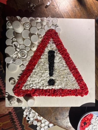 The image shows a canvas art piece depicting a red-bordered triangle with a black exclamation mark inside, resembling a warning sign. The artwork is made from recycled plastic bottle caps and other plastic fragments, predominantly in red, white, and black. The canvas is placed on a wooden surface, likely a table, with a portion of a patterned rug visible in the lower left. Scattered around the canvas are additional white plastic caps and some red and white plastic fragments in a bowl. The lighting suggests an indoor setting. There is no indication of the city of Abiansemal, Indonesia, in the image or its context.