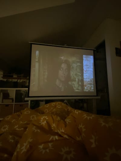 An interior scene within a residence in Boulogne-Billancourt, France, is depicted in dim lighting. A white projection screen occupies the central and upper-mid portion of the frame, displaying a video image. The projected image features a person, likely female, with dark, curly hair and an expression of concern or surprise, shown in a medium close-up against a textured background that includes a dark, possibly brick, wall on the left and a vertical section with blue light on the right. In the foreground, spanning the lower half of the image, is a bed covered with a mustard-yellow duvet or comforter patterned with white sun or starburst motifs; the bedding shows visible folds. To the left of the screen, a white multi-compartment shelving unit is partially visible, containing several light brown storage bins on its lower shelves. A dark doorway or room opening is present on the right side of the frame. No individuals are directly visible in the room, but the setup indicates a viewing activity.