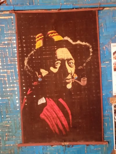 The image displays a framed, stylized portrait, likely a painting or fabric art, of a person resembling Bob Marley. The subject is depicted with dark, voluminous hair or dreadlocks, topped with a yellow and red striped hat or headwear. The face features large eyes, a prominent nose, and a beard or goatee. The individual is shown smoking a pipe and wears distinctive earrings—one blue and one orange/red. A red garment is visible around the subject's shoulders. The portrait utilizes strong lines and vibrant colors (red, yellow, white, blue) against a dark, possibly black or very dark brown, background.

The artwork is rectangular and hangs against a wall made of woven strips of material, likely bamboo or reeds, painted a bright blue. The textured, blue wall shows light spots, suggesting either light filtering through gaps or reflections, and provides a rustic backdrop. The artwork is suspended by wires or hooks attached to a visible rod at the top.

On the right side of the main artwork, against the blue wall, fragments of other papers or signs are visible. A small white label shows the word "Lear", possibly part of "Learn". The setting, with its woven blue wall, suggests an informal environment such as a market stall, a small shop, or a simple dwelling in Arambol, India. The subject's expression appears calm and contemplative.