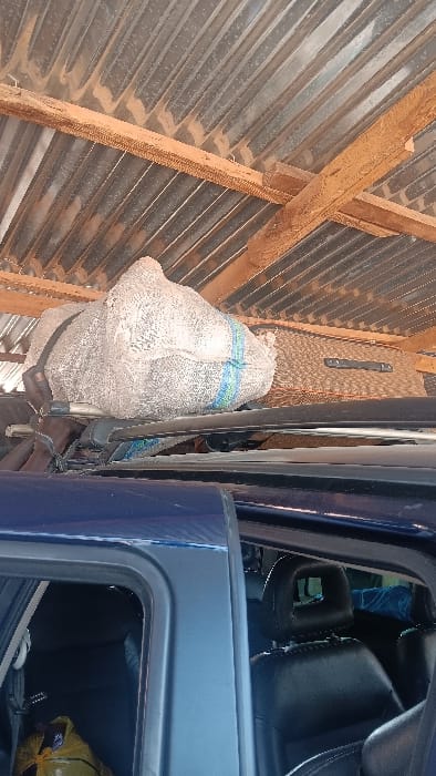 The image depicts a static scene within an open structure in Langtang, Nigeria. The upper portion of the frame shows a corrugated metal roof or ceiling, supported by wooden beams, all heavily coated with reddish-brown dust or dirt. Directly below the roof, a dark blue vehicle, likely an SUV or car, is positioned, viewed from its upper side. A roof rack is mounted on the vehicle, carrying luggage. On the roof rack, a large, bulging, light-colored mesh sack with blue and green stripes is prominent, positioned next to a brown, textured suitcase or bag. The vehicle's passenger-side door is partially open, revealing dark-colored, possibly black, car seats with headrests inside. A yellow bag is visible on the floor or seat within the vehicle's interior on the left. No individuals, actions, or interactions are visible.