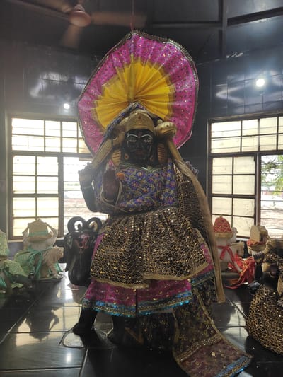 The image depicts a statue of a Hindu deity within a dimly lit room. The statue is adorned with a colorful, ornate costume featuring sequins and metallic threads. It has dark skin and is wearing a headdress that fans out like a halo. Several other smaller statues and objects are visible in the foreground and background, including what appears to be a white tiger statue and a black urn-like object with an "Om" symbol. The room has large windows with a grid pattern, suggesting they might be in a temple or shrine. A ceiling fan is visible in the upper left corner, and the floor is highly reflective, possibly made of polished tile. The lighting suggests artificial light sources within the room. There are no people visible in the image. The style of the statues and decorations is indicative of a religious or cultural site in India. While specific details about Gurgaon are not present in the visual information, such statuary is common in religious establishments throughout India.