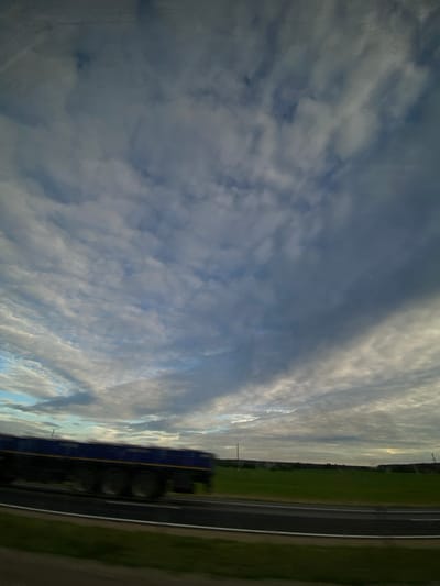 The image shows a train with a blue and yellow exterior traveling along a track. The train is positioned on the right side of the frame, with its wheels and carriages partially visible. To the left of the train, there is a vast expanse of clouds filling the sky, with hints of blue sky visible in between. The clouds are diverse, ranging from wispy, thin formations to denser, darker masses.
The scene appears to be set in a rural landscape, as indicated by the green fields stretching alongside the railway tracks. A treeline is visible in the distance, suggesting the presence of forests or a wooded area. A telephone pole stands near the train tracks, breaking the horizontal line of the horizon. The lighting suggests it is daytime, possibly late afternoon or early evening, given the interplay of light and shadow on the clouds and fields. The overall mood is serene, with the vastness of the sky and the open landscape creating a sense of tranquility. There is no visible text in the image.