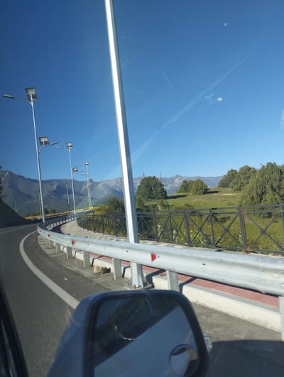The image depicts a roadside scene with a clear blue sky overhead. Several streetlights with solar panels are visible along a paved road that curves to the left. The road is bordered by a metal guardrail, behind which is a walkway with a patterned surface. Beyond the walkway, there is a grassy area with scattered trees and distant mountains. A vehicle's side mirror is visible in the lower right corner of the frame, reflecting a portion of the car's interior. There are no people visible in the scene. The location is stated to be in the city of Myk, Uzbekistan.