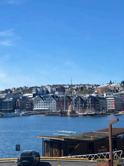 The image captures a view of Tromsø, Norway, from a waterfront perspective. The scene includes a body of water adjacent to a cityscape densely populated with buildings. A sign reads "THE HOTSPOT," indicating a commercial location. A modern building with glass accents and wooden structures sits adjacent to the waterfront. The sky is predominantly clear blue. In the lower right, a vehicle, possibly a car, is visible. The camera angle appears rotated 90 degrees. The city skyline includes a mix of residential and commercial buildings, some with distinct architectural styles and varying heights. A sailing ship mast can be seen amongst buildings.