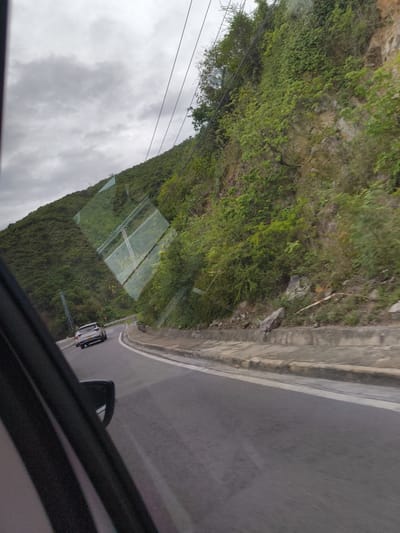 An image taken from inside a vehicle shows a gray, overcast sky above a winding asphalt road. The road is flanked by a steep, vegetated hillside. In the distance, a white SUV is driving away from the camera. Reflections on the car's window show power lines and the hillside. The road has a concrete barrier along its edge, and some debris is visible on the roadside. The scene appears to be a highway or a scenic route, possibly in a mountainous area. The location in Nam Nha Trang, Vietnam, suggests a coastal mountainous region.