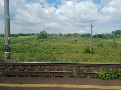 The image shows a railway track in the city of Trypillia, Ukraine, under a cloudy sky. The foreground features the railway tracks, with ballast and sleepers visible. A yellow line marks the edge of the platform. In the background, there is a vast green field with scattered trees and vegetation. Several utility poles and power lines traverse the landscape, indicating infrastructure in the area. The overall scene depicts a rural or semi-rural setting adjacent to a railway line. No people are visible in the image.