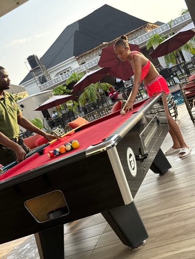 A brightly lit outdoor scene shows a woman in a red bikini leaning over a pool table, cue in hand, preparing to strike the cue ball. Her upper body is arched over the table, while her legs are angled in the opposite direction. She is wearing light-colored sandals. The pool table is red with multiple balls racked at one end. A man in a green shirt stands on the other side of the table, holding a pool cue and watching the woman. Behind the pool table, multiple tables and chairs are visible, shaded by large red umbrellas. A building with a gray tiled roof and white decorative railing stands in the background. The location is Rubuchi, Nigeria.