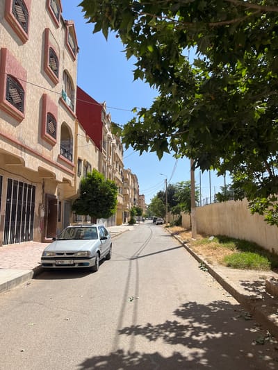 This is a vertical, eye-level, outdoor shot of a street in El Hajeb, Morocco, on a sunny day. The scene is dominated by a tall, multi-story building with a beige, textured facade and red accents, featuring numerous windows with decorative ironwork. To the left, lush green tree branches hang over the scene, casting dappled shadows. A grey sedan is parked on the right side of the paved street. The street is lined with a raised sidewalk and a retaining wall, with dry grass and some scattered debris along the edge. In the distance, the street curves and more trees are visible, with a clear blue sky overhead. The light suggests it is midday. There is some visible text on the license plate of the car, which appears to read "52700 11 61".