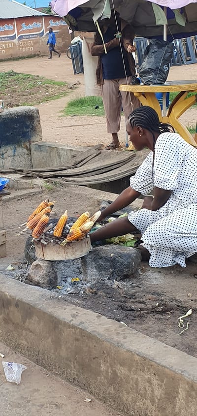 In Barakin Angundi, Nigeria, a woman with braided hair, wearing a white polka-dot dress, is shown roasting corn over an open fire. She is hunched over, tending to several cobs of corn that are arranged on a metal grill above a charcoal brazier. The brazier is made of a cylindrical object with a metal grate on top, supported by rocks. The ground around the brazier is covered in ash and dirt. In the background, other people and objects are visible. A man in a blue jacket and shorts walks away from the camera. A yellow table with a black trash bag on it is also present, as is a large umbrella providing shade. The scene appears to be an outdoor market or roadside stall.