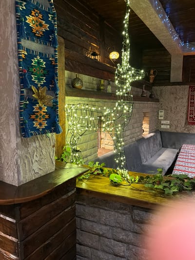 The image depicts an indoor scene with a rustic and festive ambiance, possibly in a restaurant or private residence in Kumbor, Montenegro. The central focus is a wireframe deer silhouette illuminated by numerous small, warm white LED lights. Green ivy vines are draped around the deer and the wooden ledge it stands on, adding a natural element.
To the left of the deer, a vertically hung textile tapestry with geometric patterns in blue, orange, and green is attached to a textured, pale pink wall. The tapestry features tassels and a golden leaf embellishment. Behind the deer, a brick fireplace structure is visible, with a mantelpiece displaying various decorative items, including ceramic vessels and brass pitchers.
Further in the background, a long, grey upholstered sofa is arranged along a wall. To the right of the sofa, a table is set with a tablecloth featuring red, white, and black striped patterns. The ceiling is constructed of dark wooden beams, and strings of small white and blue lights are draped along them, contributing to the festive lighting. The overall impression is one of cozy, decorated interior space with elements of traditional design.