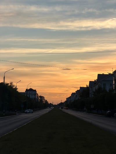 The image depicts a street scene in Dzerzhinsk, Russia, during a sunset. The sky is a gradient of pale blue and warm orange hues, with streaks of clouds. Overhead wires crisscross the frame, suggesting an urban environment. Along the right side of the road are buildings with classical architectural elements and trees lining the sidewalk. A grassy median separates the road from a sidewalk. Cars are visible on the road, indicating that this is a functional street. The setting appears to be a residential or mixed-use area. The time of day is evident from the sunset colors in the sky. The weather appears clear with scattered clouds. No people are prominently featured, and the overall mood is serene, typical of a late evening in a city. There is a yellow pedestrian crossing sign visible on the right side of the road.