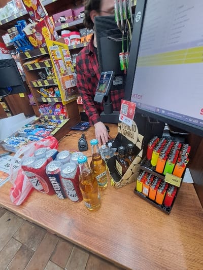 The image depicts a retail checkout counter inside a store in Bolshaya Elhovka, Russia. A male individual, wearing a red and black plaid shirt, stands behind the counter, partially obscured by a computer monitor. His right hand is visible near a black handheld barcode scanner.
The wooden counter features several items for sale or purchase. A white plastic shopping bag with red lettering is positioned on the left. Adjacent to it are eight aluminum cans of beer: four red Budweiser cans and four blue Bud Light cans, arranged in a group. To their right are three glass bottles containing a light amber liquid, possibly beer or cider, with light blue and green caps. One bottle displays the brand "ESCAPLE." Further right, a wooden-patterned cardboard box holds three dark glass bottles, likely liquor. A black display stand contains numerous disposable lighters in a variety of colors, including red, orange, yellow, and green. A white price tag displaying "49" is affixed to the lighter display.
On the counter, near the individual, is a large HP computer monitor displaying a point-of-sale interface with Cyrillic text, including "ИТОГ" (TOTAL). Below the monitor, a smartphone is mounted vertically. A red sticker with a QR code is visible on the counter.
The background consists of several shelves stocked with various retail products, including packaged snacks, candies, chips, and additional bottled goods. A yellow advertising display for "ЧИПСЫ" (chips) is prominent. The store is illuminated by overhead lighting, and the floor shows a light-colored tile pattern.