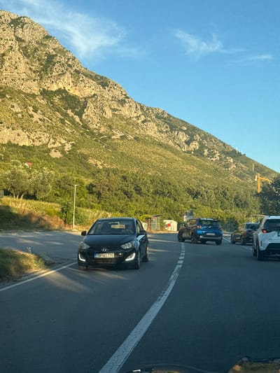 The image captures a view from a vehicle traveling on a road in Blizikuće, Montenegro. A black Hyundai i20 is in the foreground, with its license plate reading "TV AS776". Further down the road, a blue SUV is visible, followed by other vehicles. The scene is set against a backdrop of a steep, rocky mountain covered in green vegetation, with a clear blue sky above. A construction crane is also visible in the distance. The lighting suggests it is either early morning or late afternoon. There are no people visible outside the vehicles.